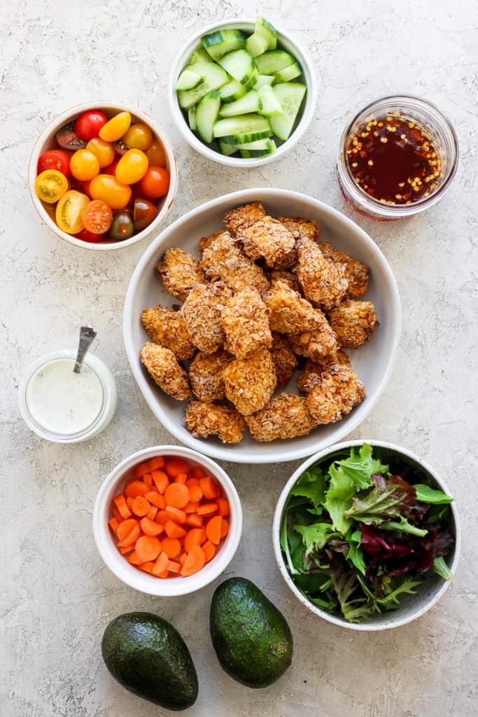 Assorted healthy food ingredients with breaded chicken pieces, bowls of fresh vegetables, mixed greens, avocado, and dips arranged on a light surface.