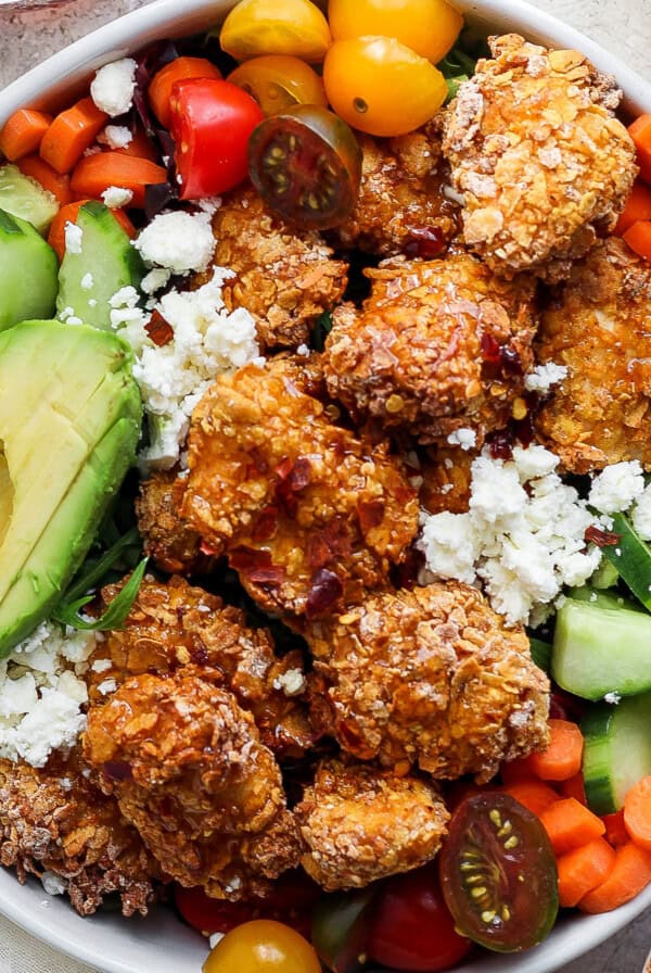 A bowl containing fried chicken, avocado slices, cherry tomatoes, sliced cucumbers, carrots, and leafy greens, topped with crumbled cheese.