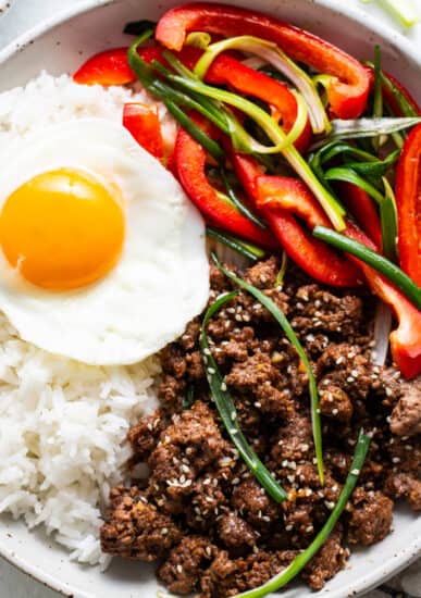A bowl of white rice topped with a sunny side up egg, seasoned Korean ground beef, sliced red bell peppers, and green onions, sprinkled with sesame seeds.