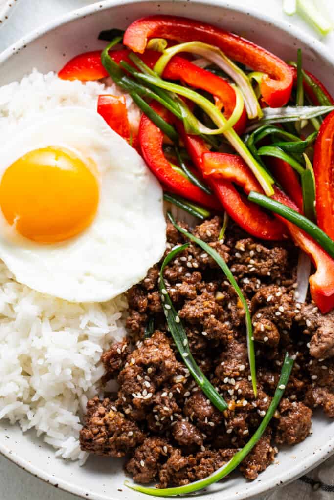 A bowl of white rice topped with a sunny side up egg, seasoned Korean ground beef, sliced red bell peppers, and green onions, sprinkled with sesame seeds.