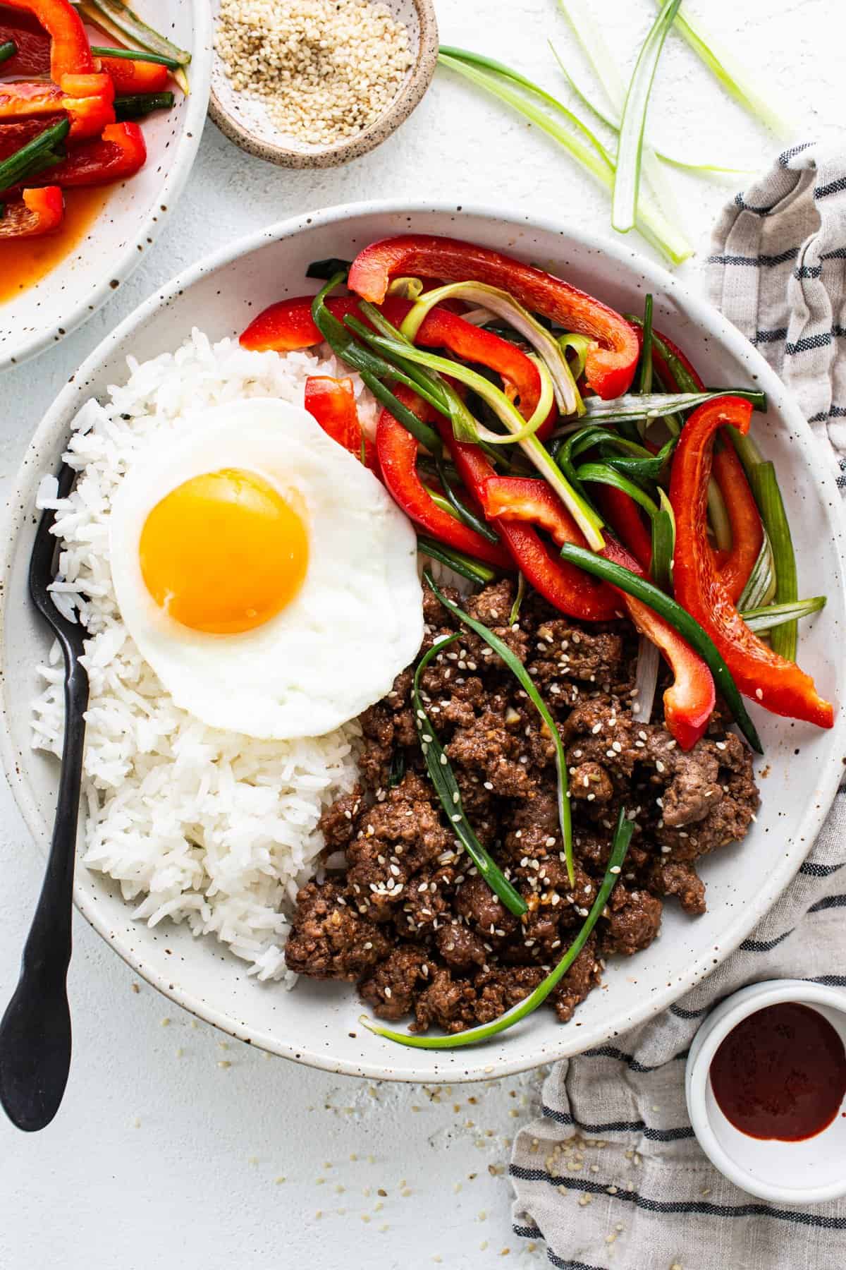 A plate of rice topped with a fried egg, stir-fried beef, and sliced bell peppers, accompanied by condiments on a white surface.