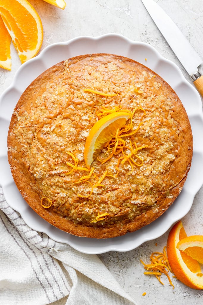 A freshly-baked orange cake garnished with orange zest on a white plate, accompanied by sliced oranges.