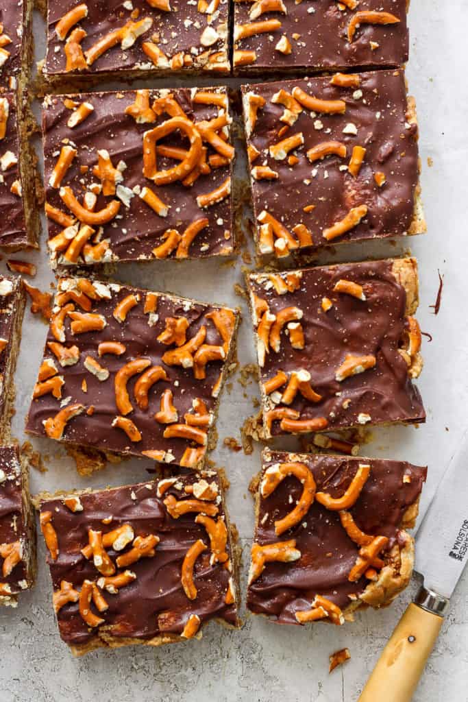 C،colate-covered toffee bars topped with pretzel pieces on parchment paper, cut into rectangles, with a knife resting to the side.