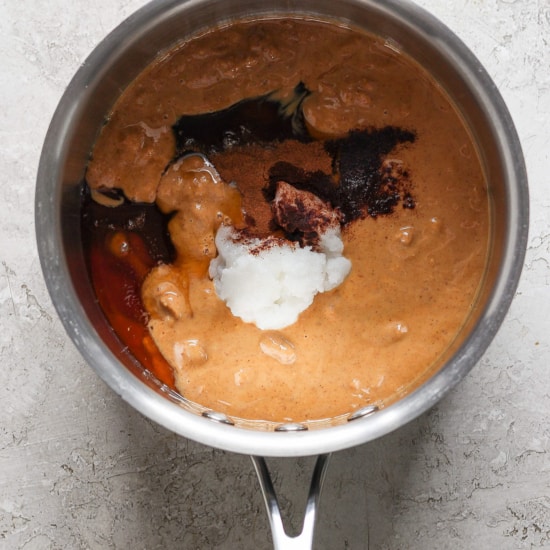 A stainless steel saucepan containing ingredients for a dish, including a reddish-brown sauce, visible spices, and a dollop of white substance, possibly coconut milk or cream, in the process of.