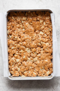 A baking tray filled with a peanut butter rice krispie treats mixture, lined with parchment paper on a light textured surface.