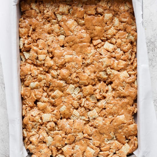 A baking tray filled with a peanut butter rice krispie treats mixture, lined with parchment paper on a light textured surface.