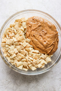 A glass bowl containing a mixture of cereal pieces and a large dollop of peanut butter on a light surface.