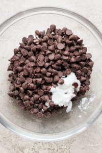 A bowl containing chocolate chips with a dollop of what appears to be white ingredient, possibly a dairy product such as cream or yogurt, on top, set against a neutral background.