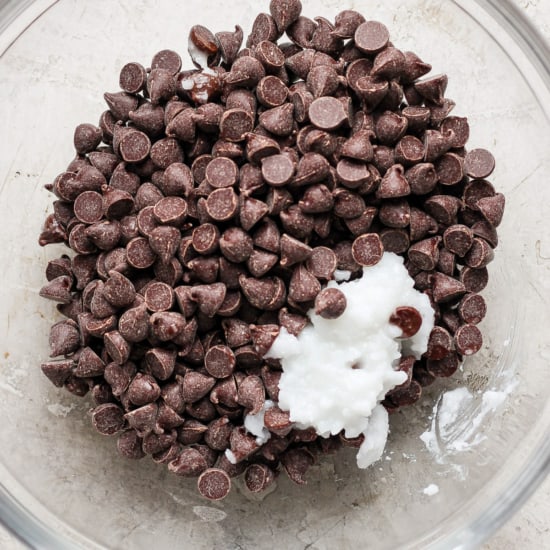 A bowl containing chocolate chips with a dollop of what appears to be white ingredient, possibly a dairy product such as cream or yogurt, on top, set against a neutral background.