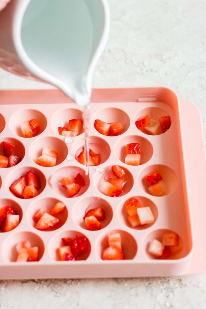 Pouring liquid into an ice cube tray filled with diced strawberries.