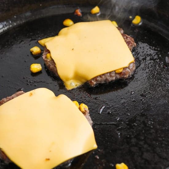 Two beef patties with melted cheese slices on top cooking in a skillet.