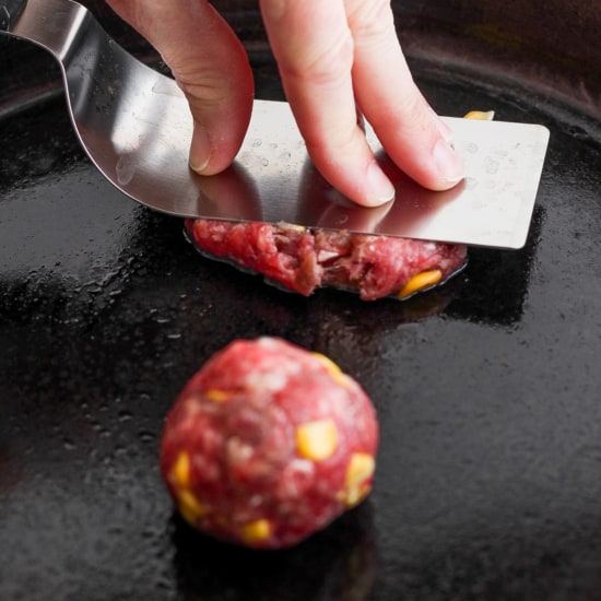 A person flattening a ball of ground meat on a skillet to form a patty.