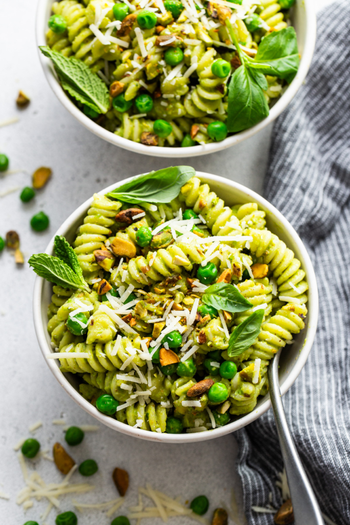 A bowl of pesto pasta garnished with peas, nuts, basil leaves, and grated cheese, served on a light surface.