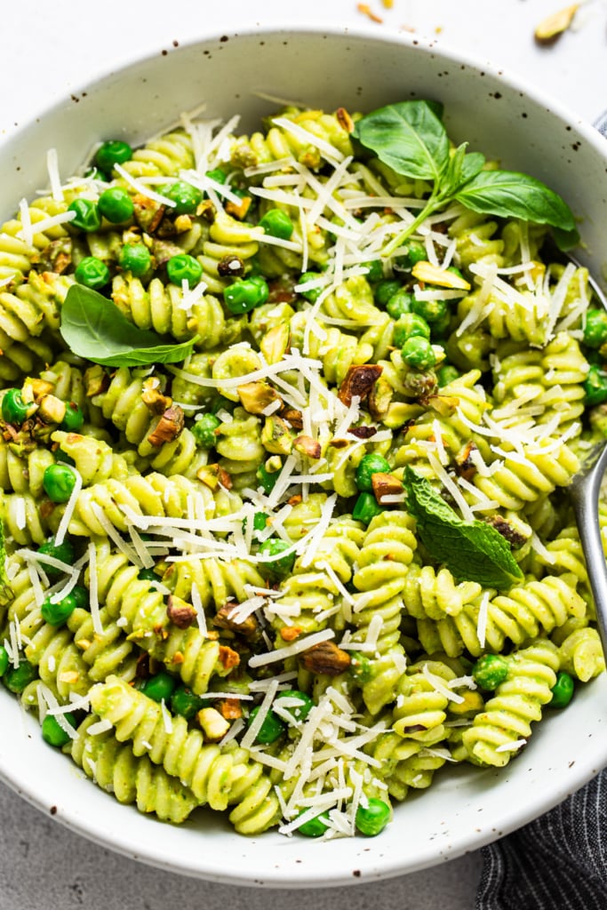 A bowl of pesto pasta with peas and grated cheese, garnished with herbs.