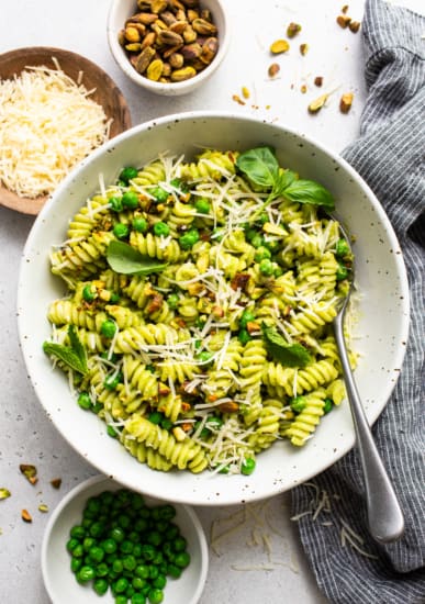 A bowl of pesto pasta garnished with grated cheese and nuts, with ingredients like peas and basil leaves on the side.