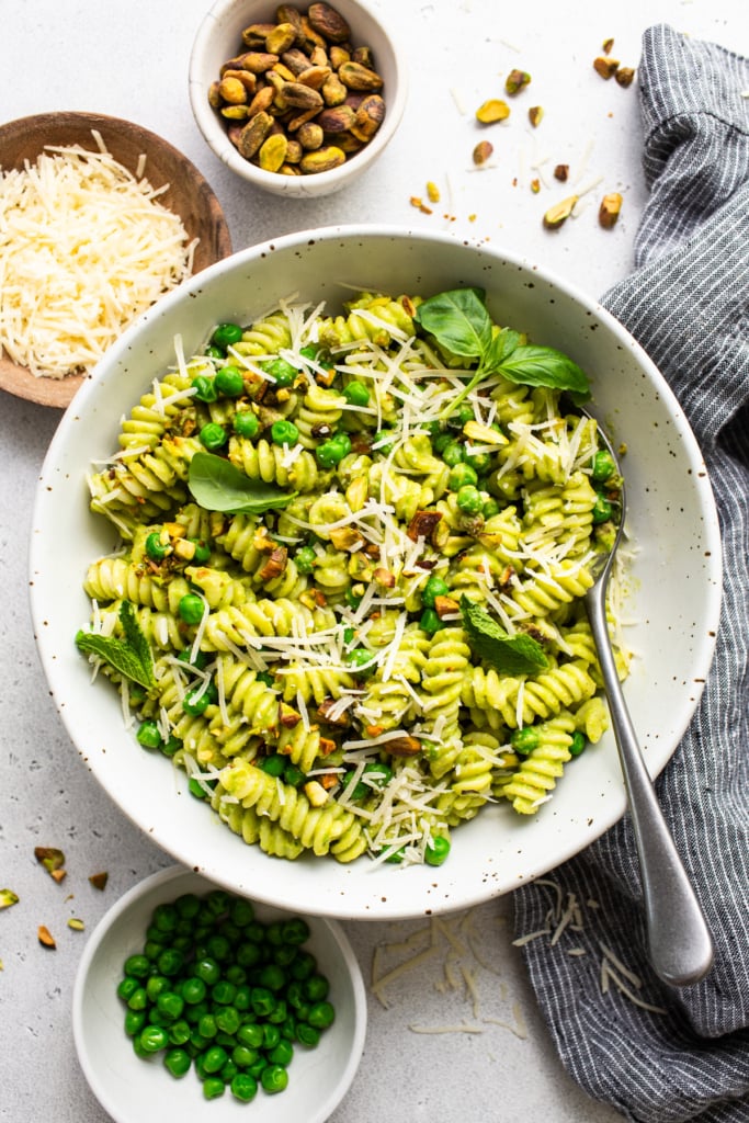 A bowl of pesto pasta garnished with grated cheese and nuts, with ingredients like peas and basil leaves on the side.