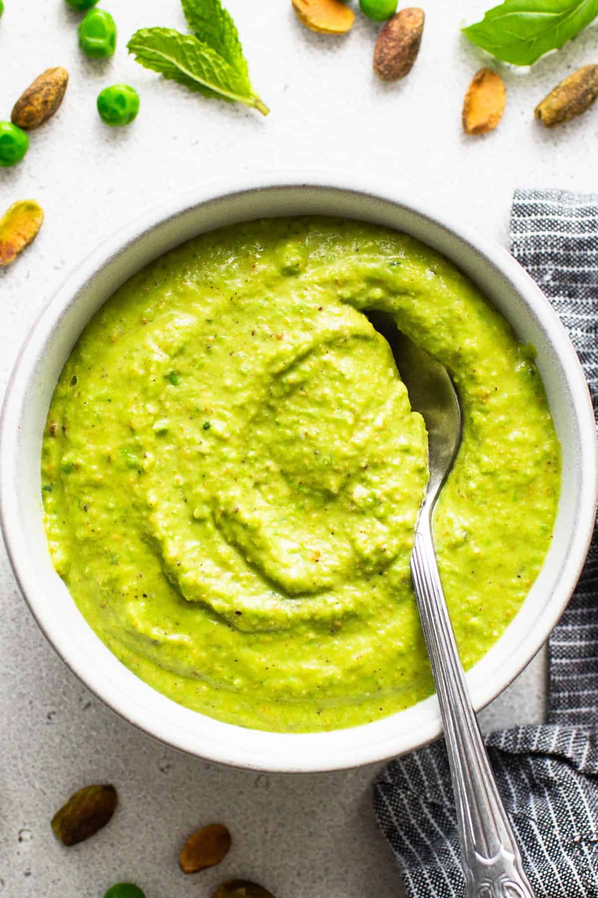 A bowl of creamy green pea and mint soup with a spoon, surrounded by peas, mint leaves, and spices on a light surface.