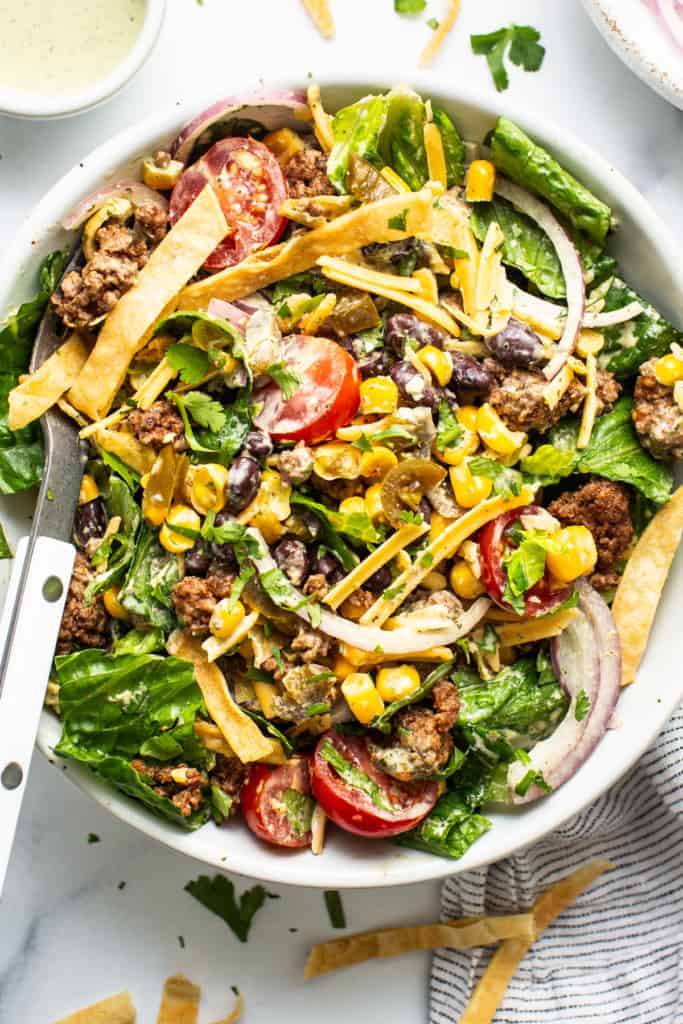 A vibrant taco salad with ground meat, mixed greens, tomatoes, cheese, corn, black beans, and crispy tortilla strips, served with a creamy dressing on the side.