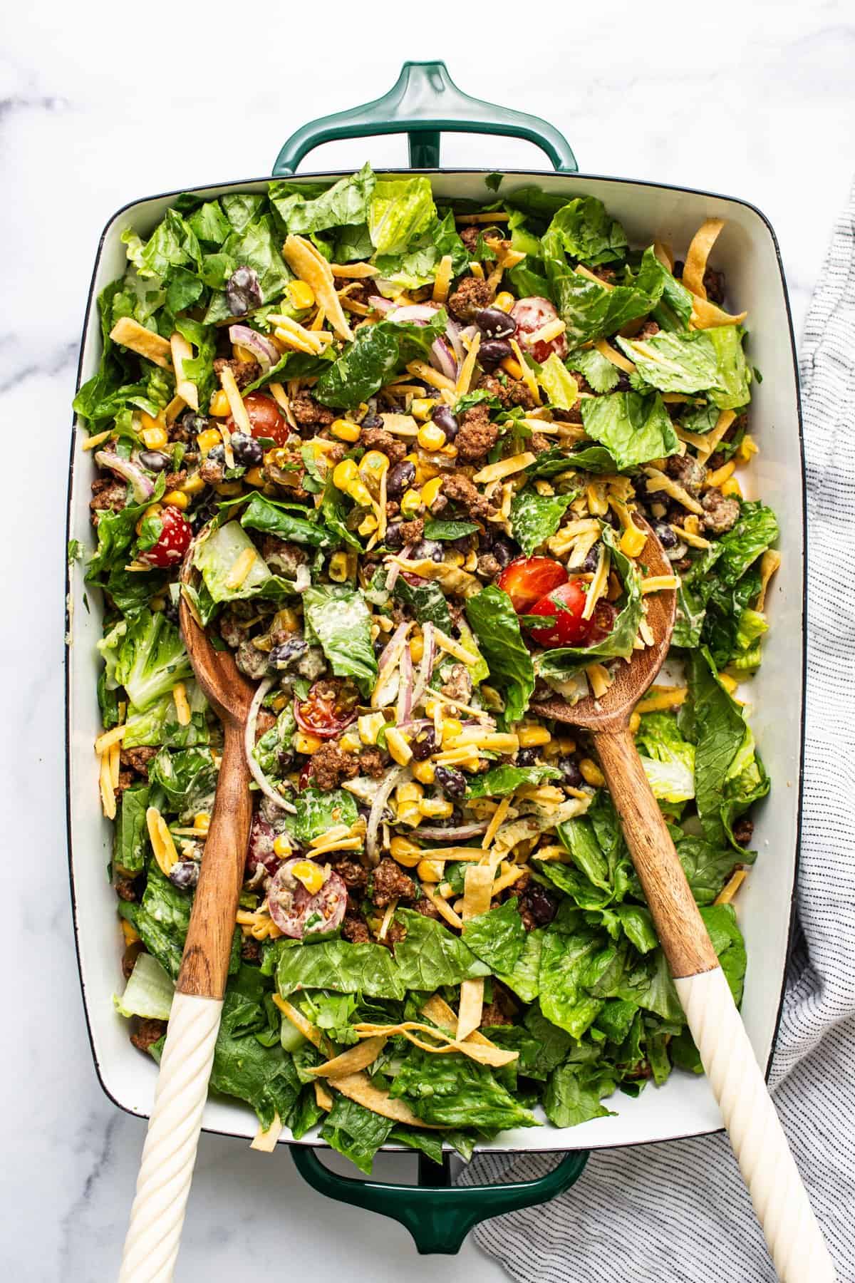 A colorful mixed salad with various greens, nuts, cheese, and tomatoes served in a large green dish with wooden serving utensils.