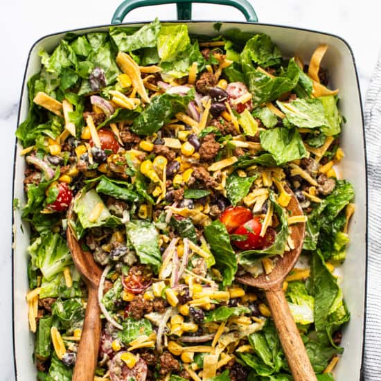 A colorful taco salad with lettuce, tomatoes, ground beef, cheese, and black beans served in a rectangular dish with wooden serving utensils.