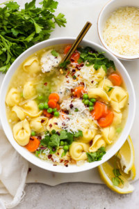 A bowl of chicken noodle soup garnished with parsley, shredded cheese, and a sprinkle of red pepper flakes, served with lemon wedges on the side.