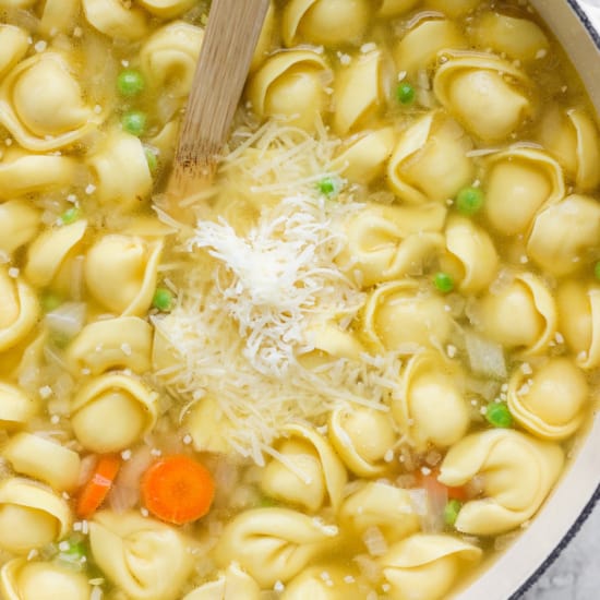 A pot of tortellini soup with carrots, peas, and grated cheese on top.