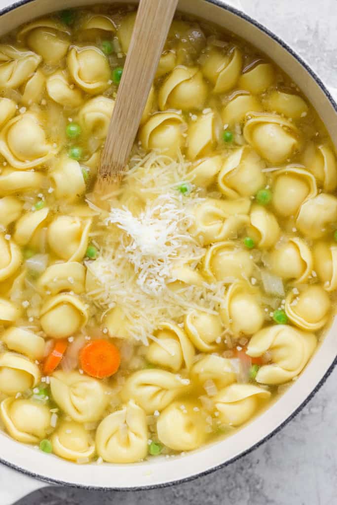 A pot of tortellini soup with carrots, peas, and grated cheese on top.