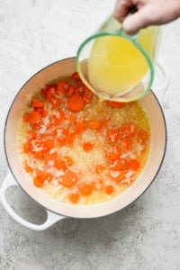 Pouring chicken broth into a pot with cooking onions and carrots.