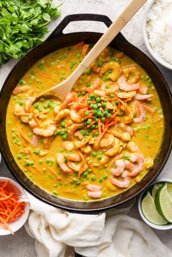 A skillet filled with shrimp and vegetable curry, garnished with green peas and served with sides of rice, lime, and fresh herbs.