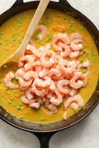 A skillet filled with a creamy pea soup topped with a mound of cooked shrimp, with a wooden spoon resting inside the pan.