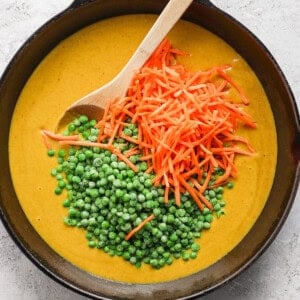 A cast iron skillet containing creamy soup garnished with a spoonful of green peas and a scattering of grated carrots on a light-colored surface.