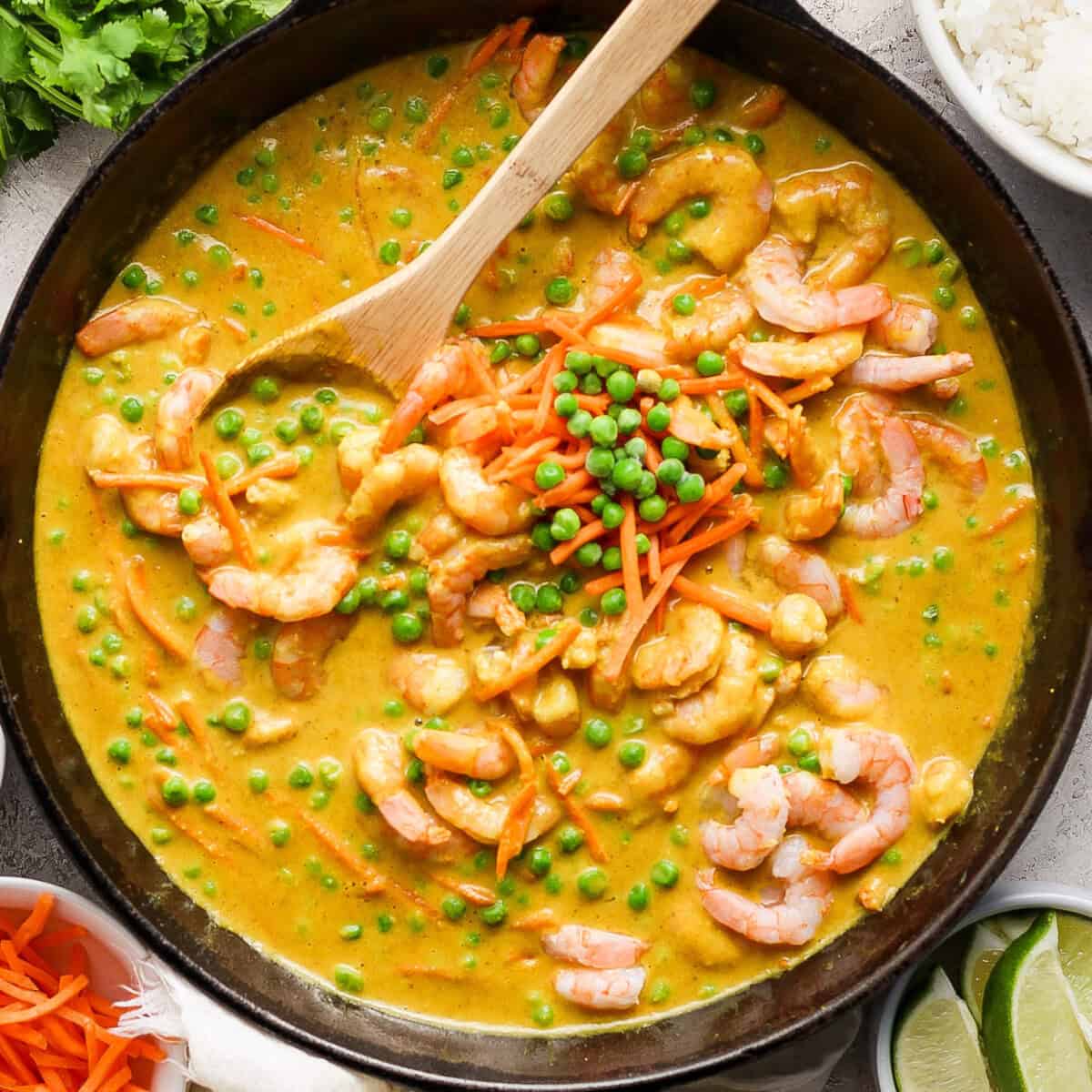 A wooden spoon is stirred into a skillet containing a shrimp curry with green peas and shredded carrots, accompanied by a side of white rice and lime wedges.