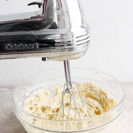 A person is using a silver cuisinart hand mixer to blend batter in a clear glass bowl on a kitchen countertop.