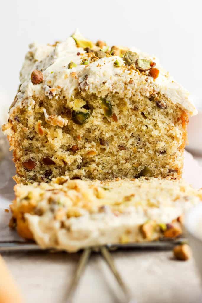 A close-up of a slice of pistachio cake with frosting and chopped nuts on top, with the remaining cake in the background.