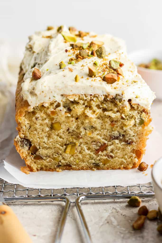 A loaf of frosted cake with nuts and chopped pistachios on top, partially sliced, displayed on a piece of parchment paper.