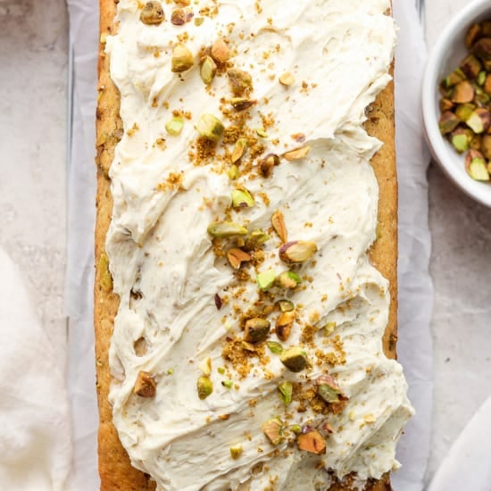 A freshly baked loaf cake topped with a creamy white frosting and sprinkled with chopped pistachios.