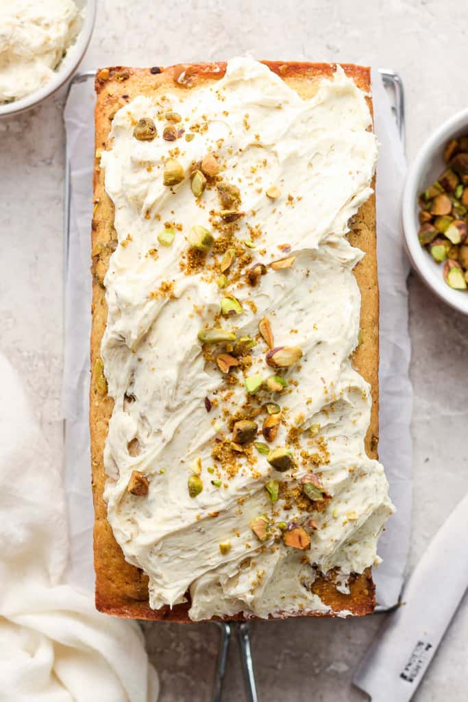 A freshly baked loaf cake topped with a creamy white frosting and sprinkled with chopped pistachios.