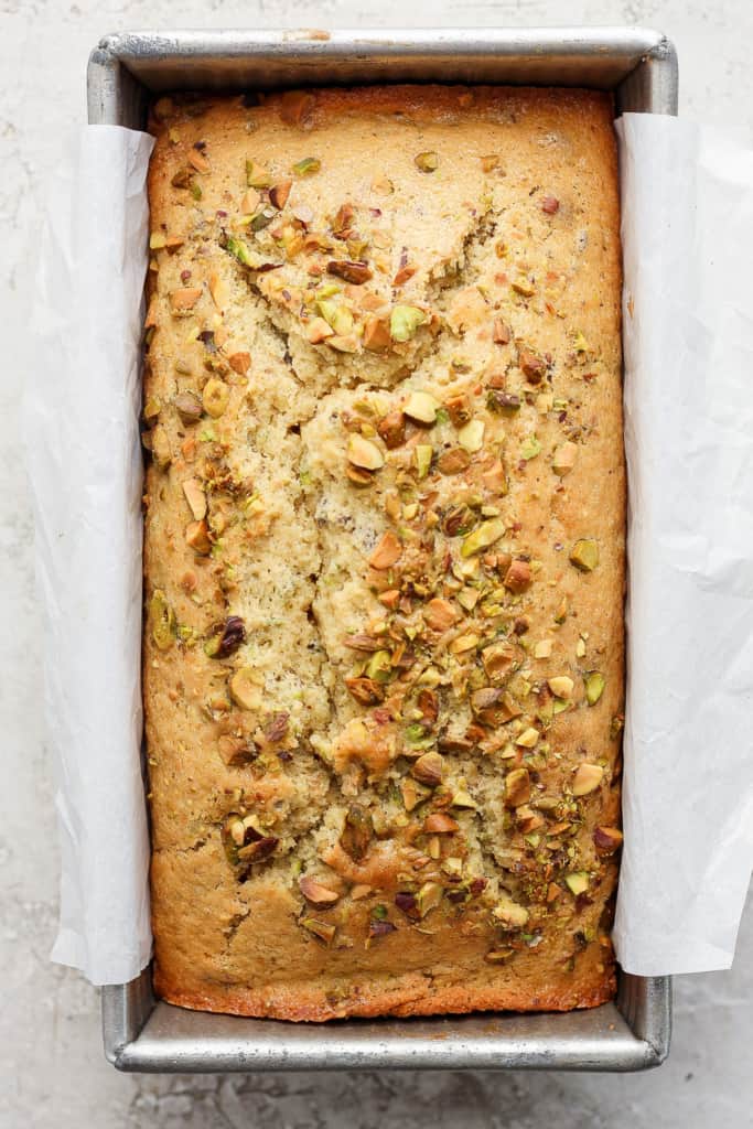 A freshly-baked loaf of bread with pistachios sprinkled on top, sitting in a metal baking pan lined with parchment paper.