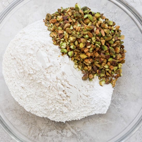 A clear glass bowl containing a pile of white flour and shelled green pistachios on a neutral surface.