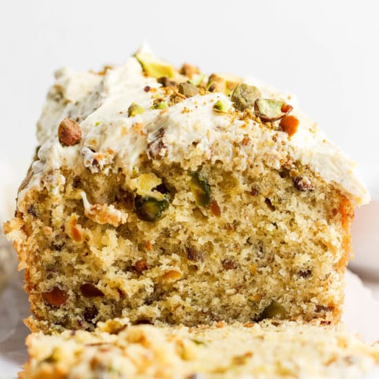 A close-up image of a sliced pistachio cake with creamy frosting and crushed nuts on top.