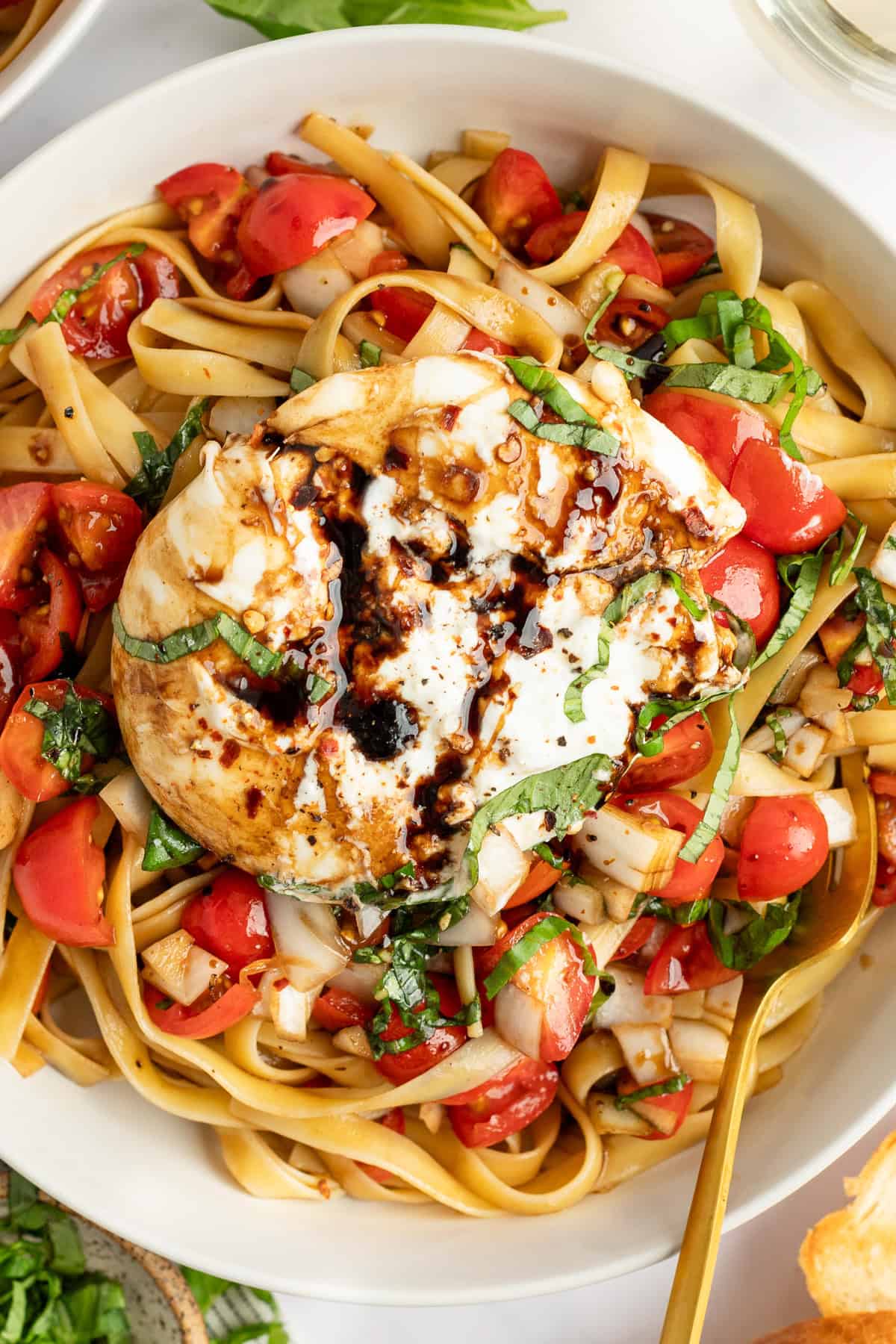 A dish of pasta topped with a large burrata cheese, drizzled with balsamic glaze, and garnished with chopped tomatoes and basil.