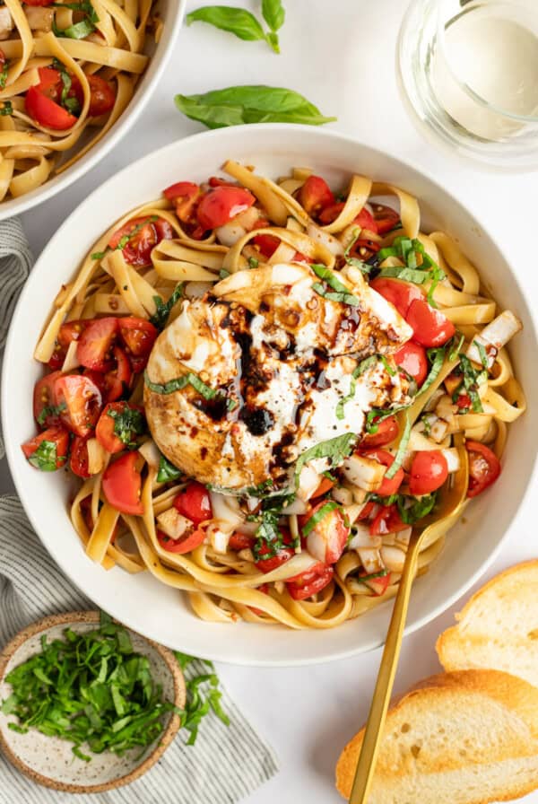 A bowl of pasta with tomatoes, basil, and a large ball of burrata cheese, drizzled with balsamic glaze, accompanied by slices of bread.