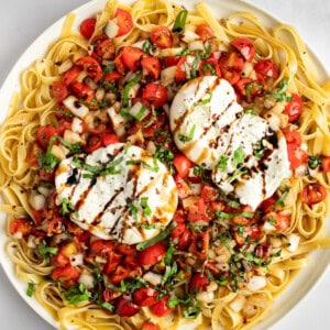 A plate of pasta topped with two poached eggs, cherry tomatoes, herbs, and a drizzle of balsamic glaze.