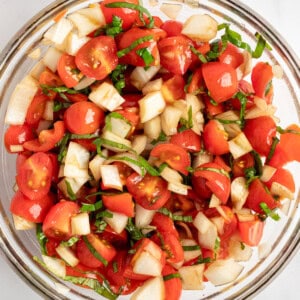 A glass bowl filled with a fresh salad of chopped tomatoes, cucumbers, and herbs on a white surface.