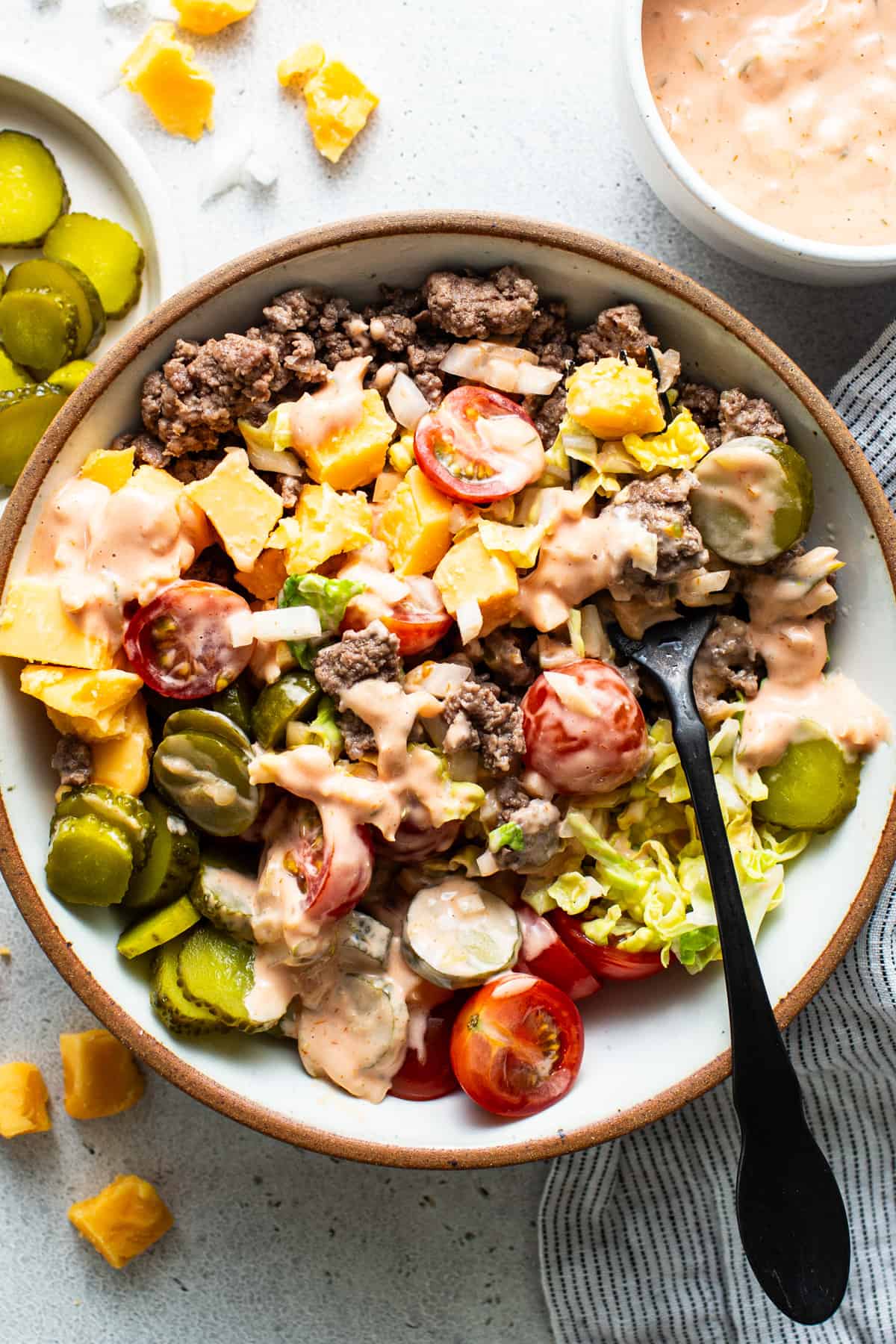 A bowl of salad with ground beef, tomatoes, pickles, cheese, and lettuce drizzled with dressing, next to a dipping sauce.