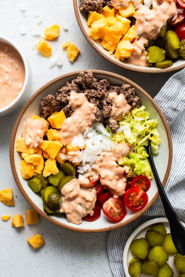 A bowl of salad with ground beef, rice, lettuce, tomatoes, pickles, olives, and diced mango, topped with a creamy dressing.