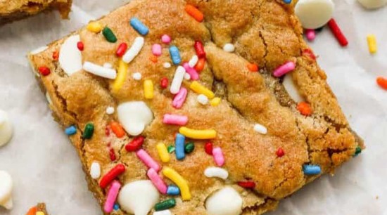 A close-up of a square Funfetti blondie topped with colorful sprinkles and white chocolate chips, with more scattered around it.