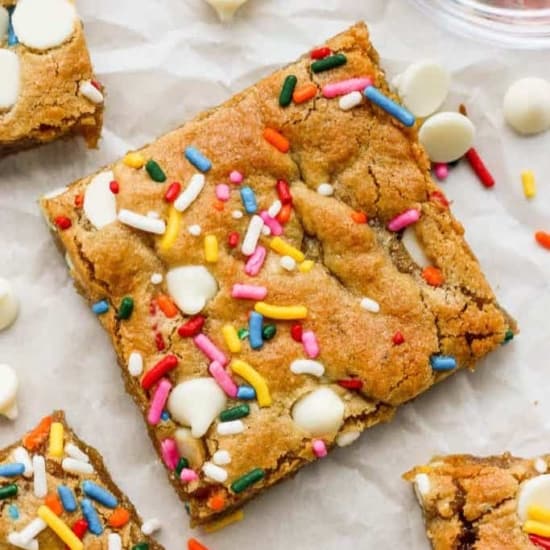A close-up of a square Funfetti blondie topped with colorful sprinkles and white c،colate chips, with more ،tered around it.