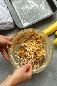 Person mixing banana baked oatmeal batter in a glass bowl next to a baking tray and fresh bananas on a gray counter.