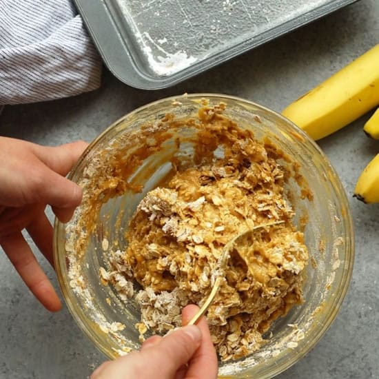 Person mixing banana baked oatmeal batter in a gl، bowl next to a baking tray and fresh bananas on a gray counter.